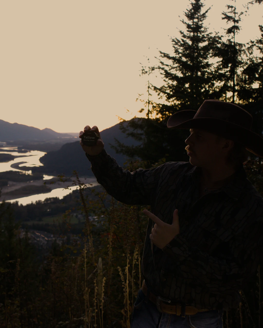 A man in a cowboy hat holding a jar of Tameco tallow shaving cream at sunset overlooking a forested valley